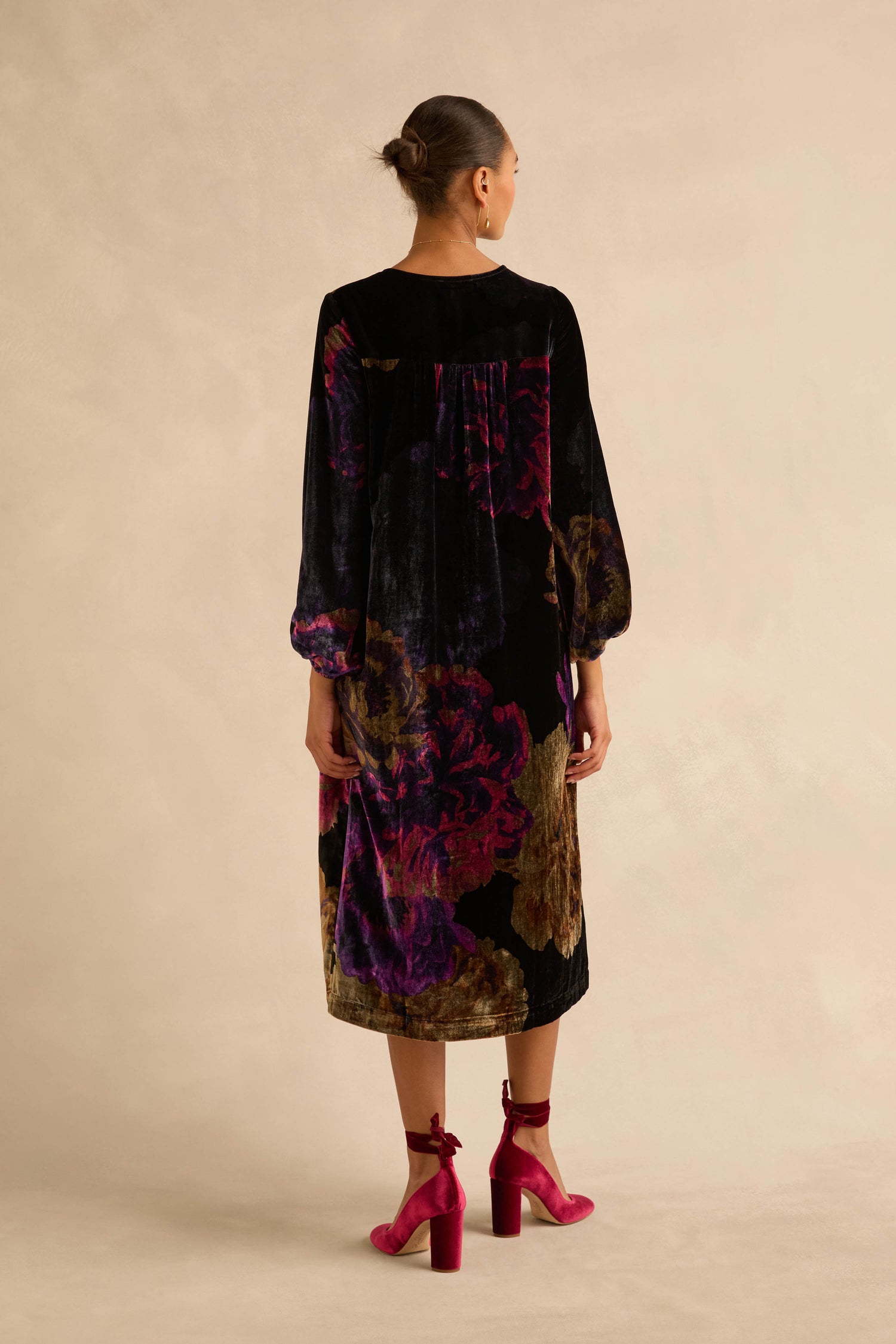 A woman wears a Midnight Peonies Velvet Dress, a long-sleeved midi style with a photographic floral print, paired with fuchsia block-heeled shoes.