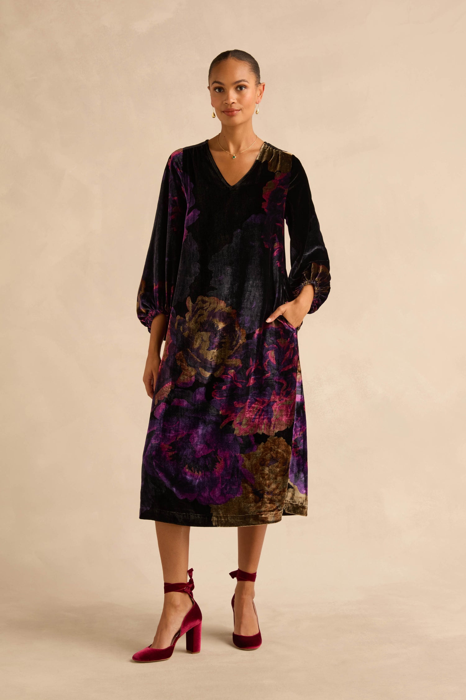 A person wears the Midnight Peonies Velvet Dress, a midi length, V-neck, long-sleeved dress with a dark purple and black floral print and pockets, paired with maroon ankle-strap block heels.