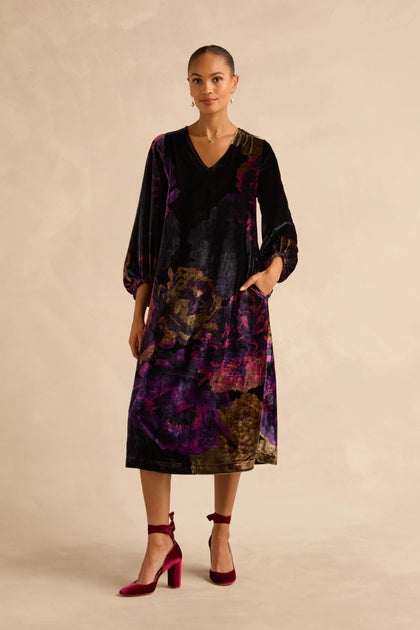 A person wears the Midnight Peonies Velvet Dress, a midi length, V-neck, long-sleeved dress with a dark purple and black floral print and pockets, paired with maroon ankle-strap block heels.