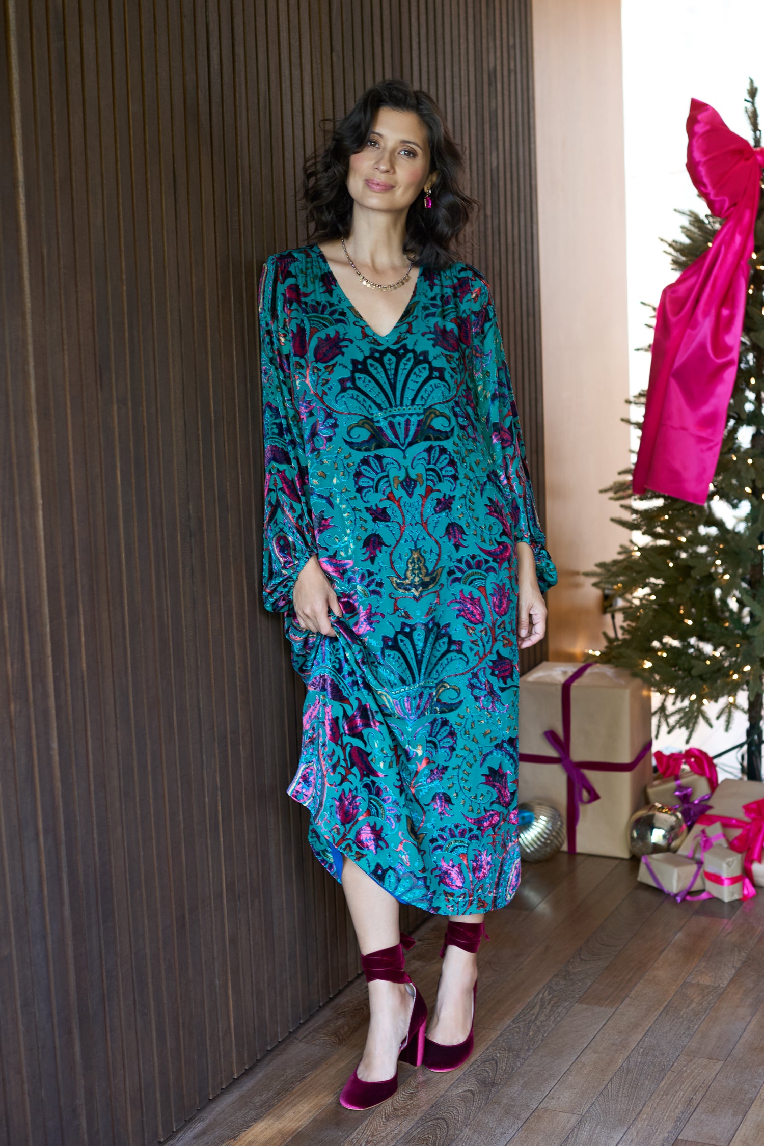 A woman wearing the Winter Paisley Devore Dress stands indoors beside a dark wooden wall and a decorated Christmas tree with wrapped gifts.