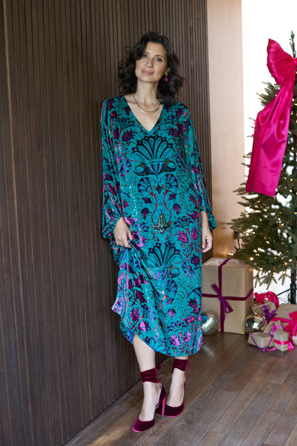 A woman wearing the Winter Paisley Devore Dress stands indoors beside a dark wooden wall and a decorated Christmas tree with wrapped gifts.