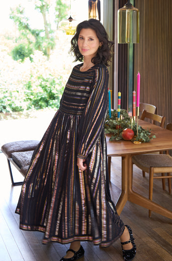A woman in the Multi Stripe Lurex Dress stands indoors near a wooden dining table decorated with candles and greenery, sunlight streaming through large windows.