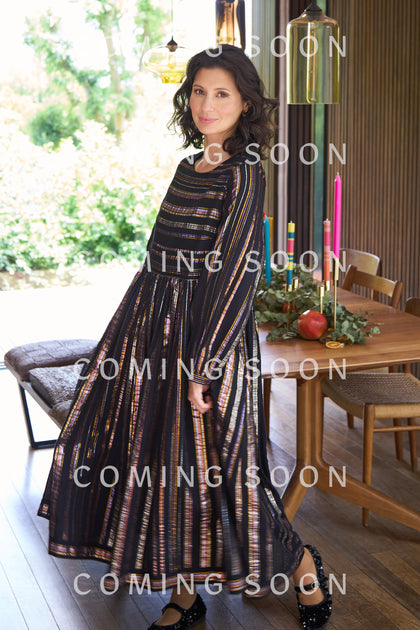 A woman in a Multi Stripe Lurex Dress stands indoors by a dining table adorned with colorful candles and fruit, with "Coming Soon" displayed over the image.