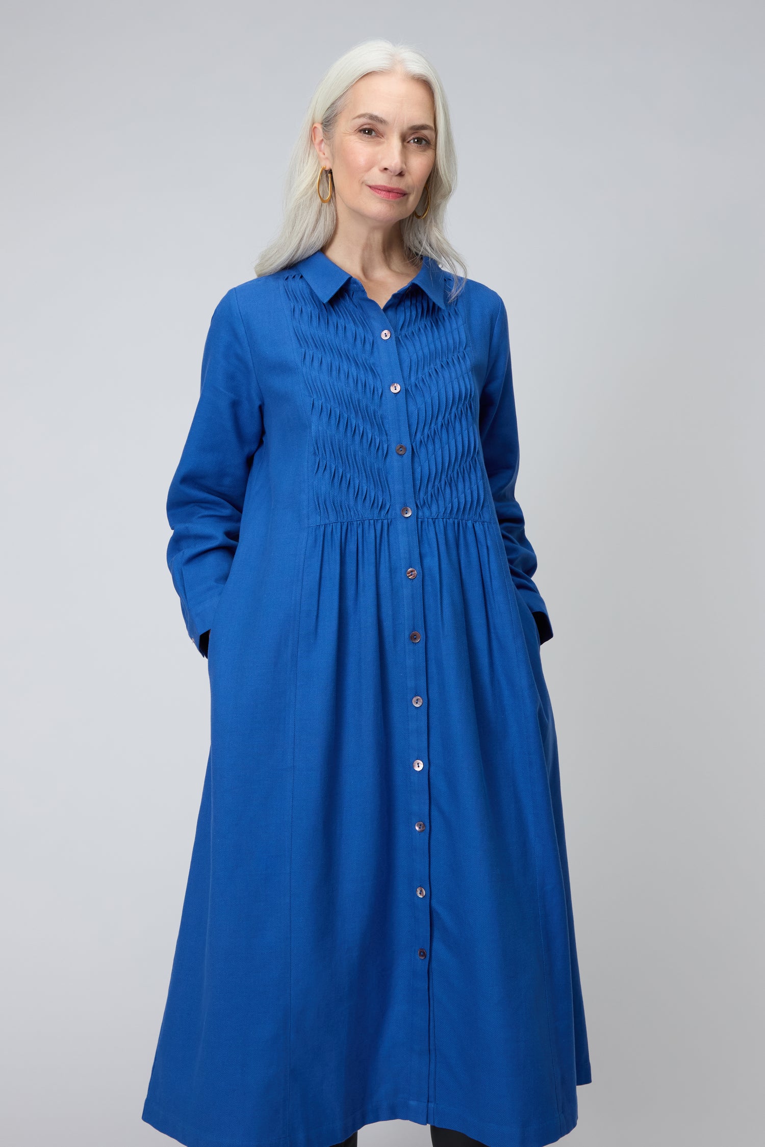 An older woman with long gray hair stands facing forward, wearing a bright blue Brushed Cotton Pintuck Detail A-line Dress with pintuck detailing on the bodice and long sleeves.