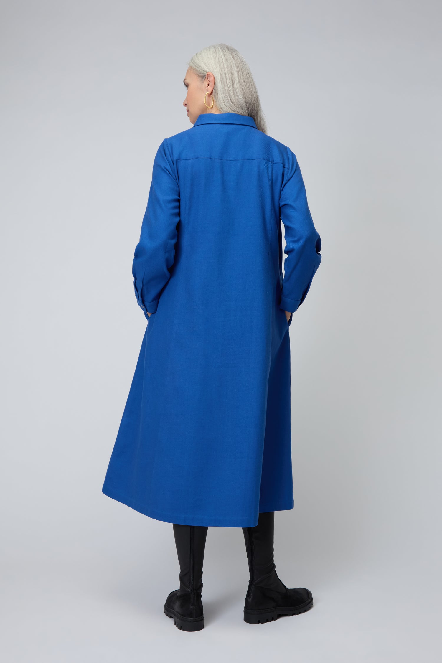 A person with long gray hair, dressed in the Brushed Cotton Pintuck Detail A-line Dress and black boots, stands facing away against a plain light background.