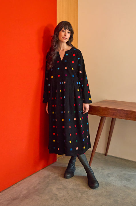 A woman in the Multi Colour Check Cord Print Dress stands by a wooden table against a red and beige wall on a concrete floor.
