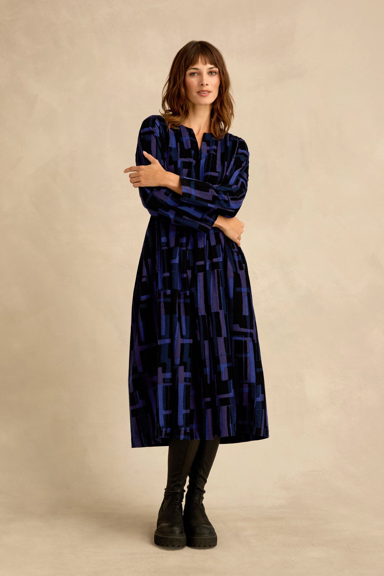 A woman stands against a neutral background, wearing the Layered Geometric Cord Flared Dress— a long-sleeve, knee-length blue and black piece featuring geometric patterns—paired with black leggings and black boots.