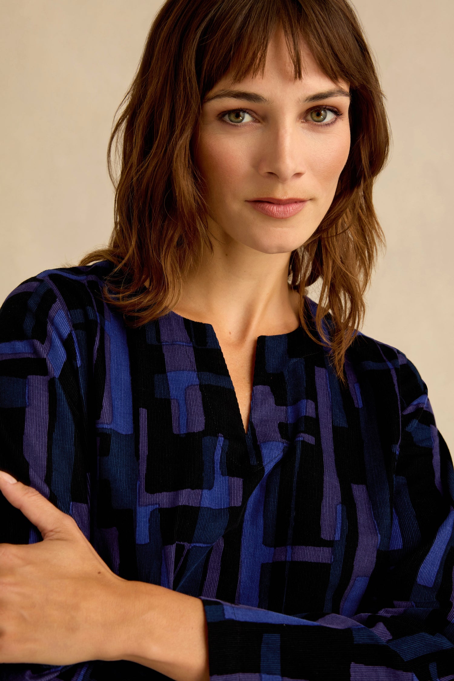 A woman with shoulder-length brown hair stands with her arms crossed, looking at the camera, wearing the Layered Geometric Cord Flared Dress in blue and black cotton needlecord.