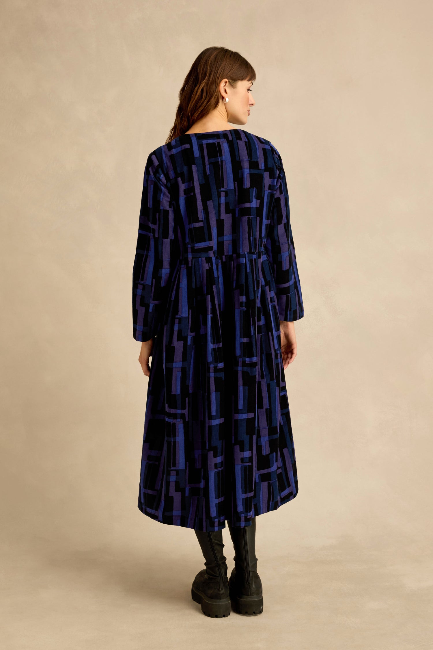 A woman stands with her back to the camera, wearing the Layered Geometric Cord Flared Dress in blue and black with long sleeves, paired with black boots, against a beige background.