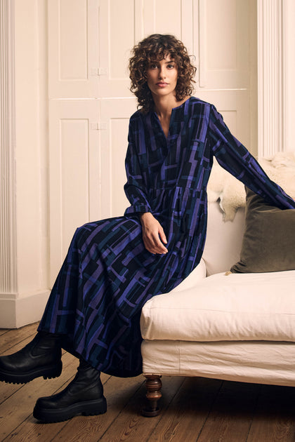 A woman wearing a Layered Geometric Fine Cord Flared Dress with a blue and purple grid pattern sits on a white sofa, paired with black ankle boots.