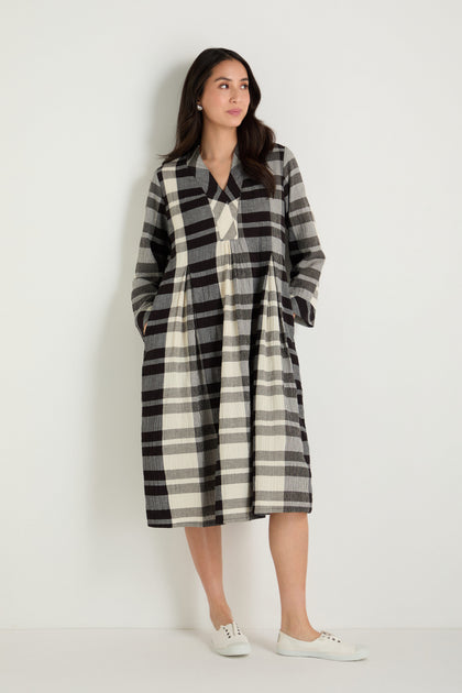 A woman stands against a white background wearing the Monochrome Crinkle Check Dress, a long-sleeved, ankle-length dress with a bold black, white, and gray plaid pattern and GRD6796-MOCC black slip-on shoes.