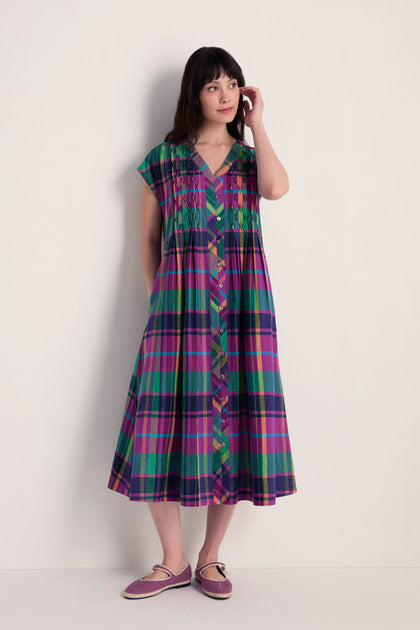 A woman stands against a plain wall wearing the Madras Check Dress with short sleeves and a button-down front, paired with matching flat shoes.