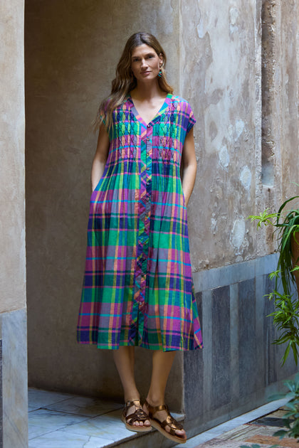 A woman stands in a hallway wearing the Madras Check Dress—a colorful cotton midi with front buttons and pockets—paired with brown sandals. The background includes textured walls and tiled flooring.