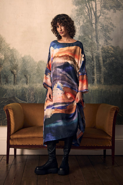 A person wearing a Moontide Silk Relaxed Dress in a colorful landscape print with black platform boots stands in front of a couch.