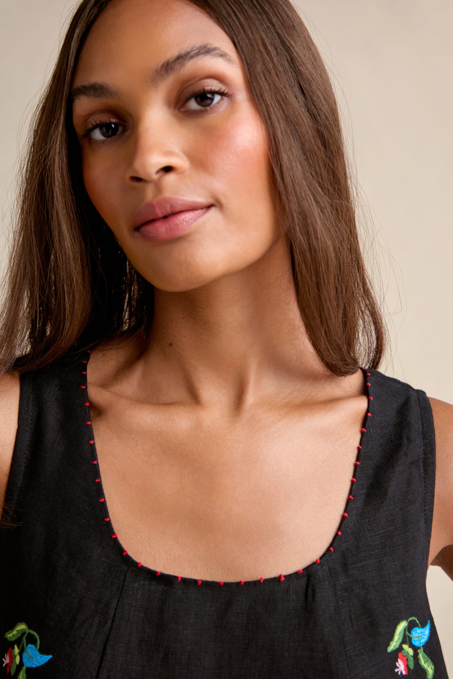 A woman with long brown hair wears the Vintage Floral Embroidered Bubble Dress, a sleeveless black top with a square neckline, red stitching, and small blue and green floral embroidery for a vintage-inspired look.