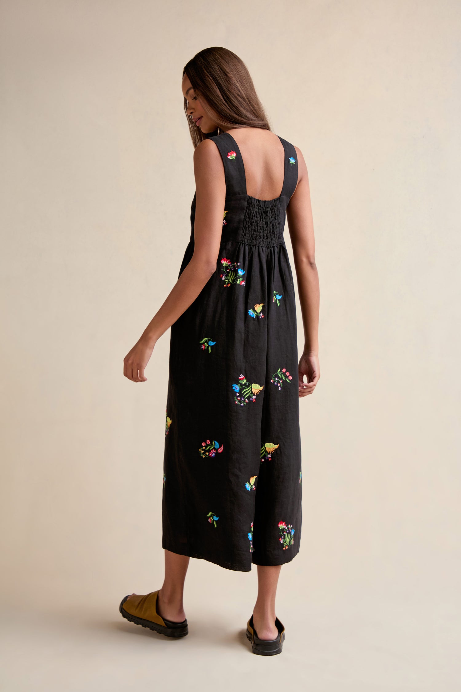 A woman stands with her back to the camera on a neutral background, modeling the Vintage Floral Embroidered Jumpsuit—a black, sleeveless piece featuring colorful floral embroidery and a back-smocked design.