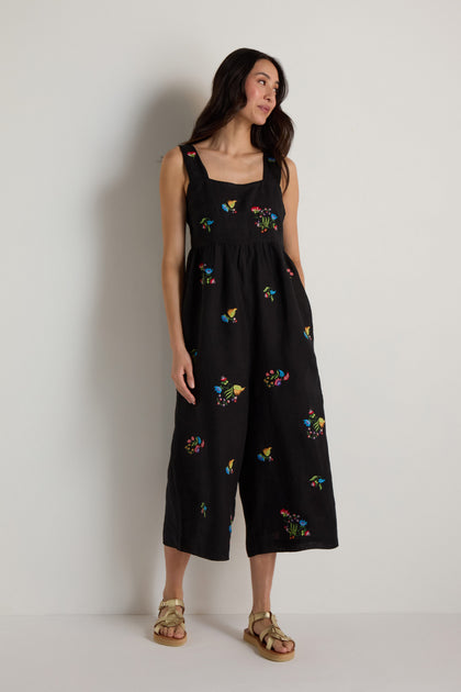 A woman models the Vintage Floral Embroidered Jumpsuit, featuring sleeveless black fabric with colorful embroidery, as she stands against a plain white wall and gazes to the side.