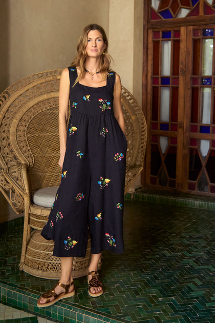 A woman stands indoors on green tiles, wearing the Vintage Floral Embroidered Jumpsuit, brown sandals, and a necklace beside a wicker chair and a wooden stained-glass door.