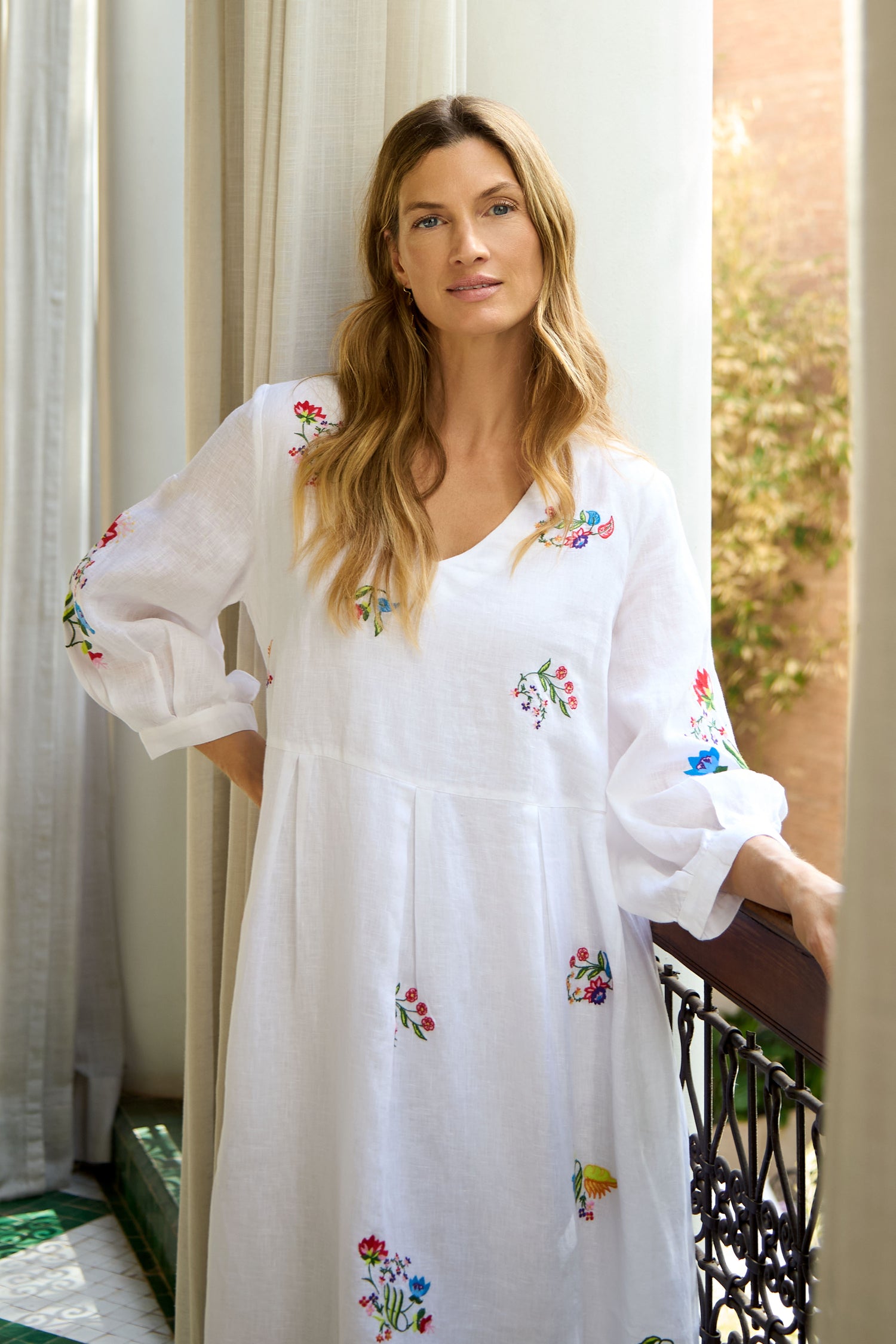 A woman with long blonde hair wears the Vintage Floral Embroidered Flare Dress, standing on a balcony with one hand on the railing, surrounded by flowing curtains and lush greenery.