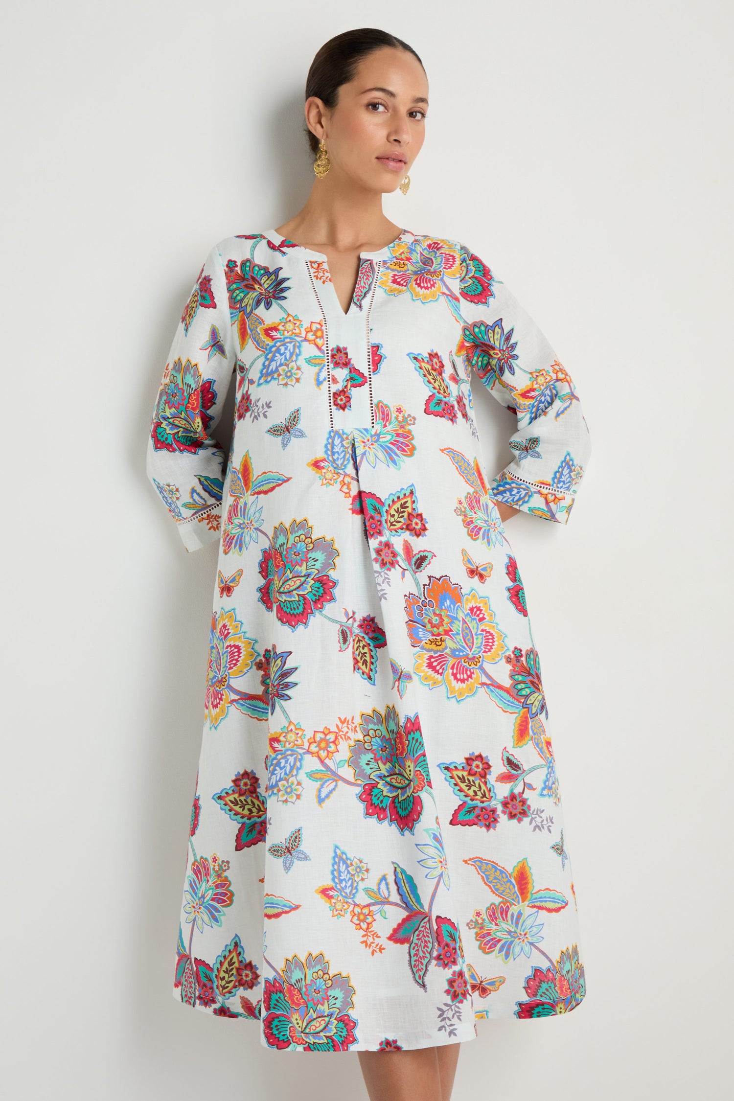 A woman models the Botanical Linen Flare Dress, a long-sleeved white linen dress adorned with vibrant botanical prints, standing before a light background.