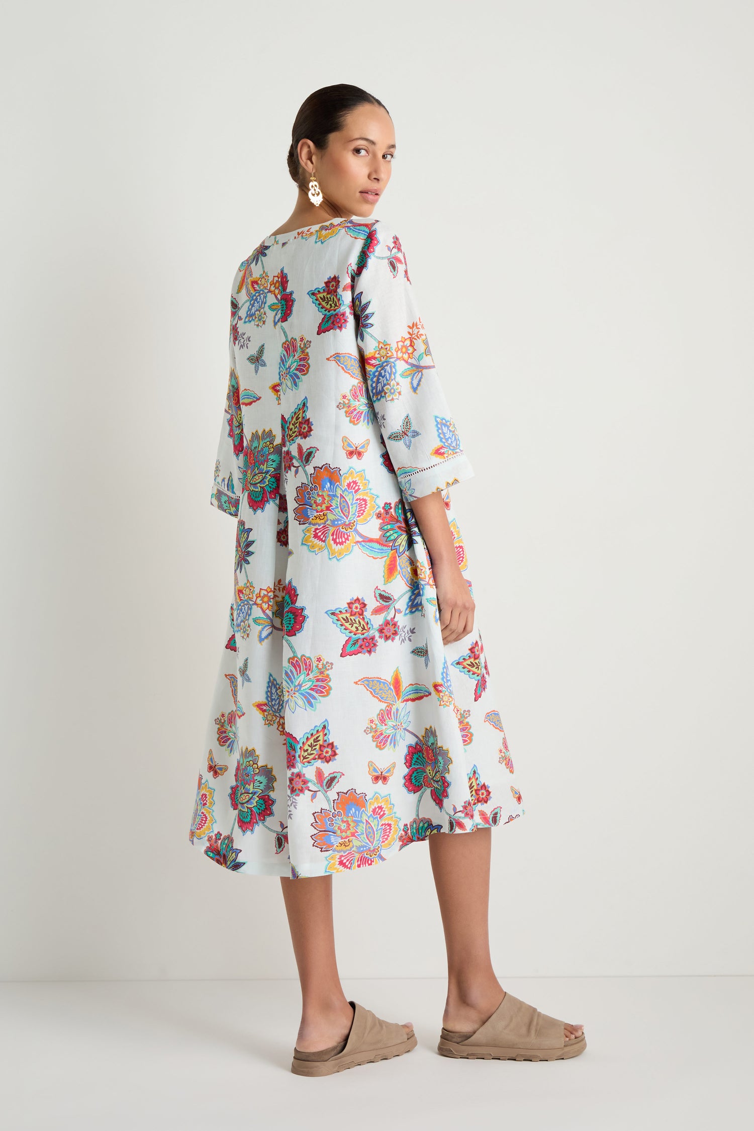 A woman stands in profile against a plain white background, wearing the Botanical Linen Flare Dress with a vivid botanical print, paired with beige sandals and large earrings.