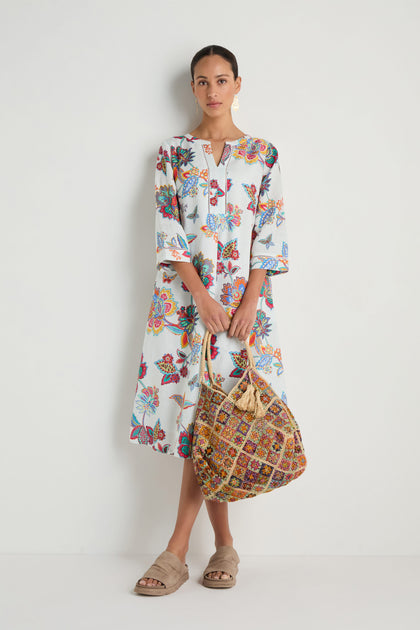 A woman stands against a white wall wearing the Botanical Linen Flare Dress, featuring a floral print, and holds a large woven bag with beige slide sandals.
