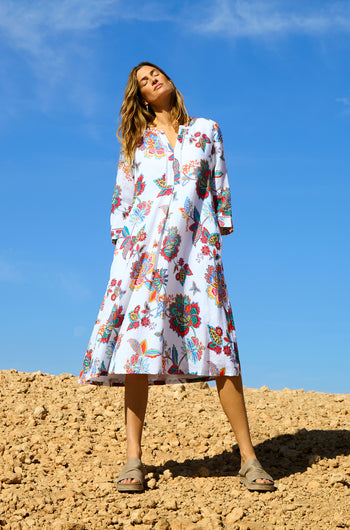 A woman stands outdoors on sandy ground under a clear blue sky, wearing the Botanical Linen Flare Dress with a colorful floral pattern and beige sandals.