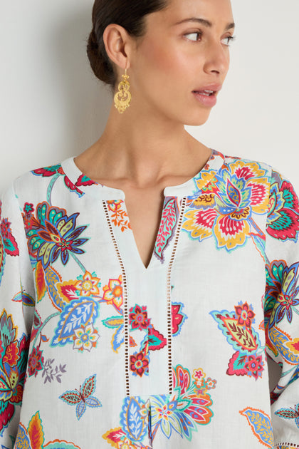 Woman wearing the Botanical Linen Flare Dress with colorful floral patterns and gold earrings, standing against a light background and looking to the side.