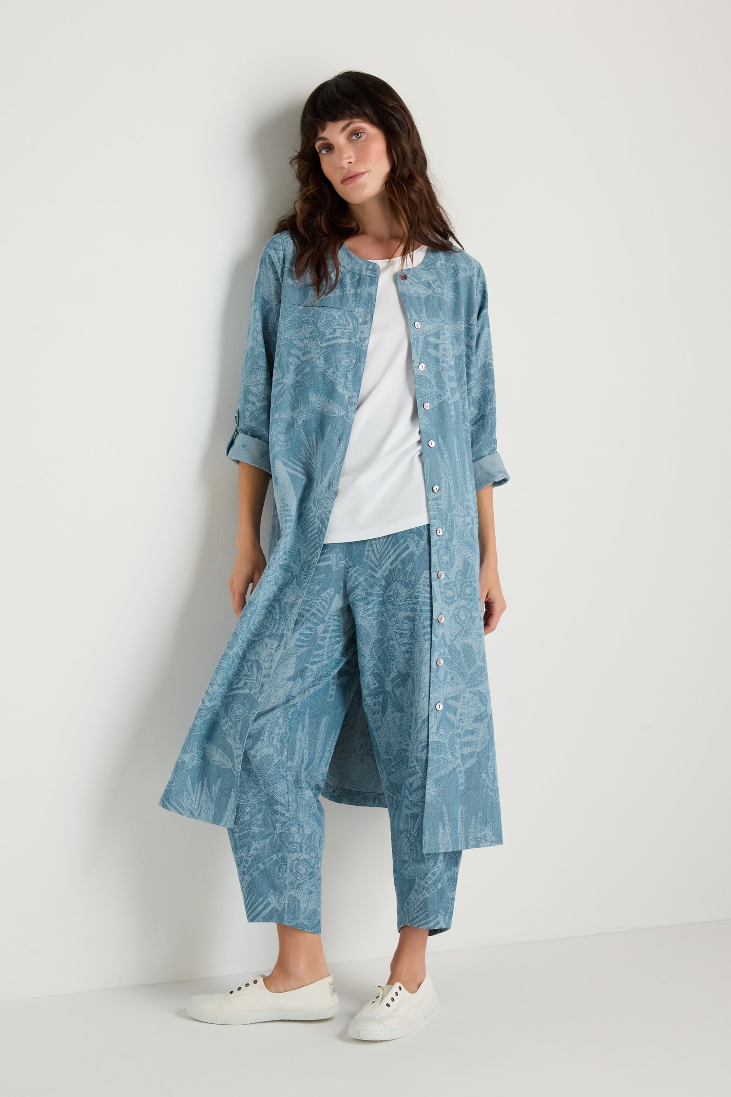 A woman stands against a plain white wall in a blue patterned Floral Jacquard Cotton Shirt Dress over a white shirt, paired with matching pants and white sneakers.