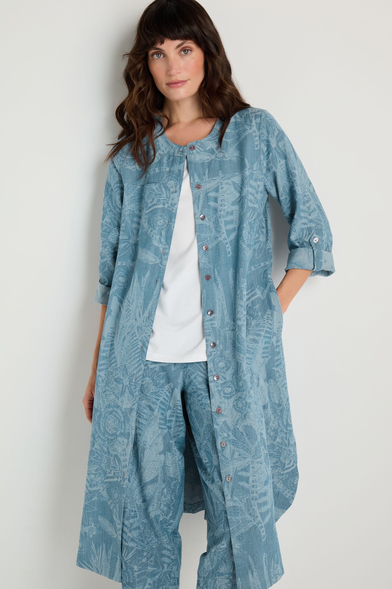 A woman stands against a plain wall wearing the Floral Jacquard Cotton Shirt Dress with rolled sleeves layered over a white top and matching blue pants.