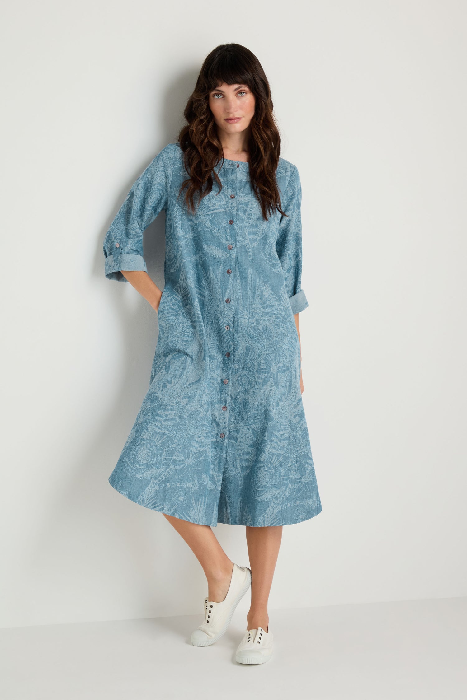 A woman in the Floral Jacquard Cotton Shirt Dress with a subtle light blue floral pattern and rolled-up sleeves pairs it with white sneakers, standing against a plain white wall.