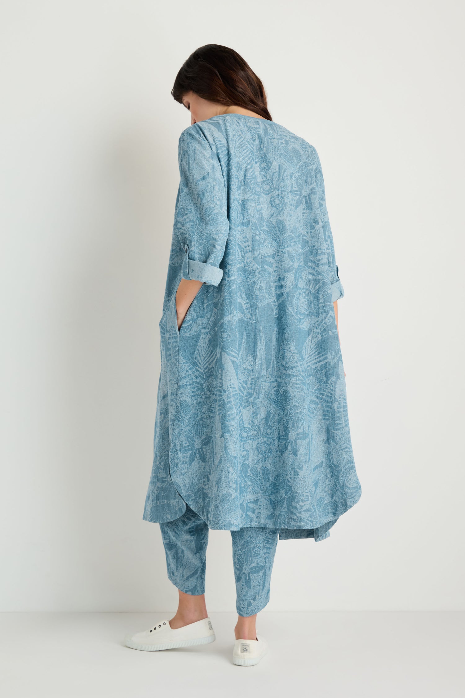 A person stands with their back to the camera, wearing the Floral Jacquard Cotton Shirt Dress in light blue with rolled sleeves and matching trousers, paired with white slip-on shoes.