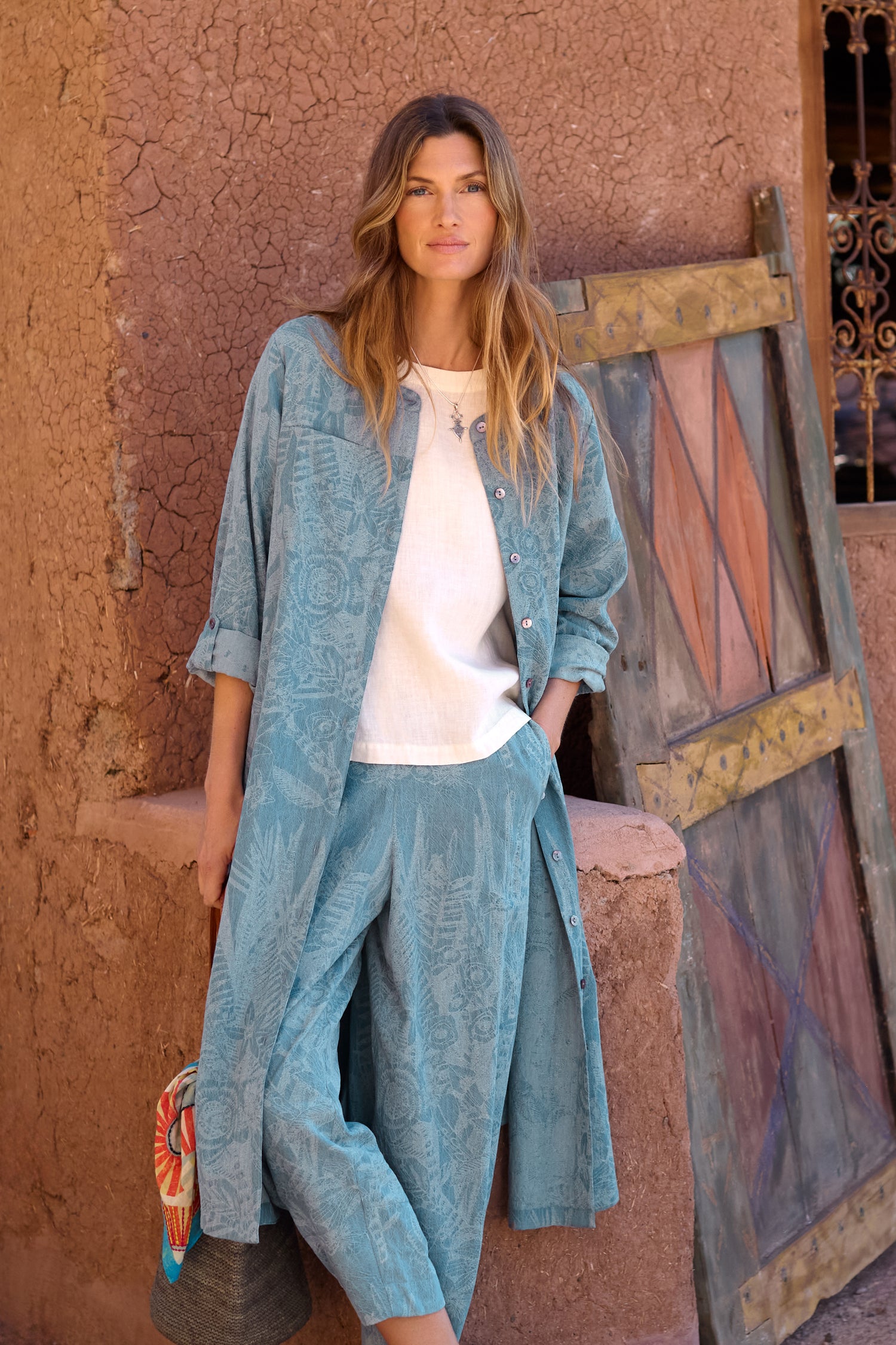 A woman wearing the Floral Jacquard Cotton Shirt Dress (style code GRD6854-FJC) stands by a rustic wall and wooden door, holding a bag and looking at the camera.