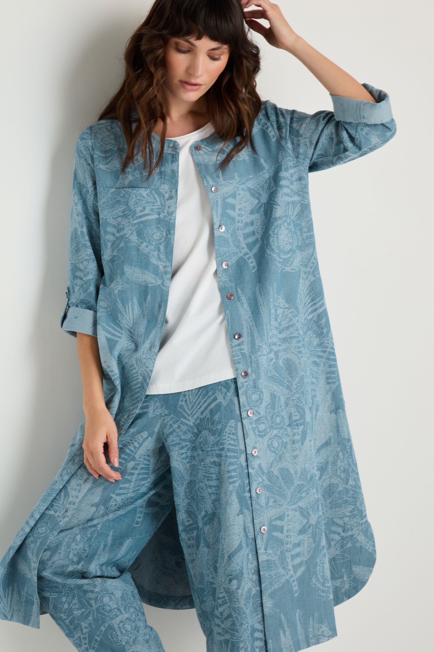 A woman wears the Floral Jacquard Cotton Shirt Dress in blue over a white top and matching blue pants, standing against a plain white background.