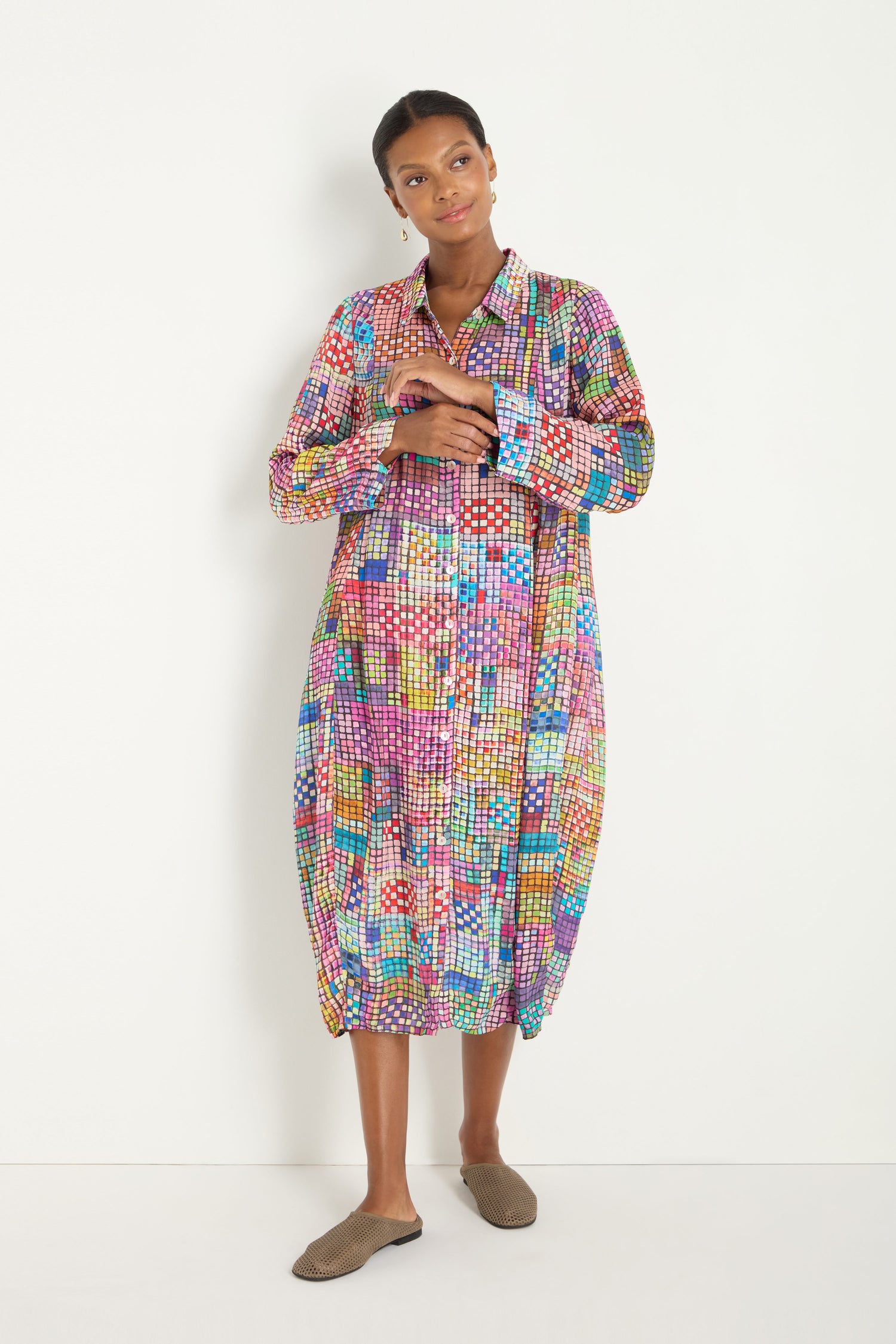 A woman stands against a white wall wearing the Coloured Glass Tiles Shirt Dress, featuring loose long sleeves and geometric patterns inspired by Moroccan mosaics, paired with black strappy heels.