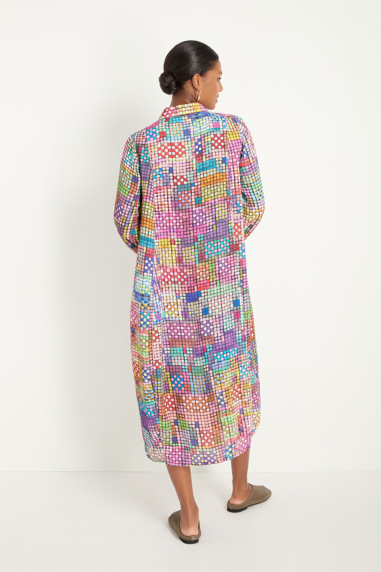 A woman stands in profile against a plain white background, wearing the Coloured Glass Tiles Shirt Dress—a long dress with a vibrant geometric mosaic print—and black strappy heels.