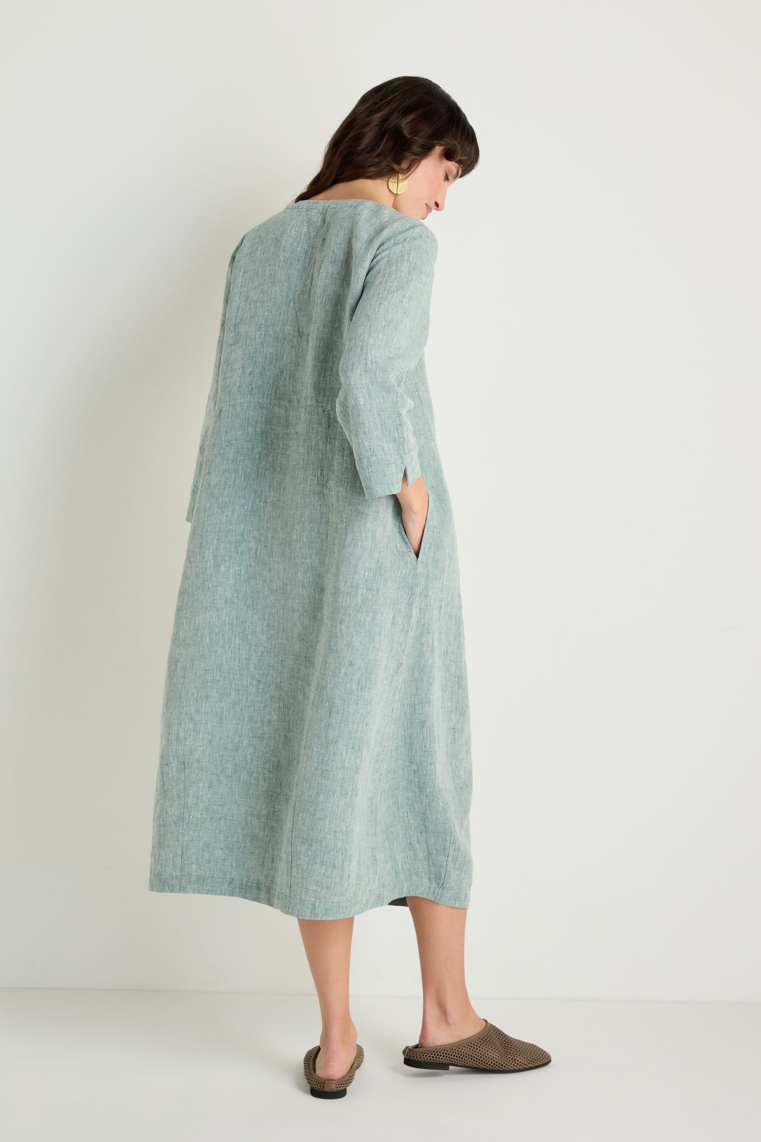 A woman faces away against a white background wearing the Crinkle Linen Cross Dye Bubble Dress, a loose-fitting, long light blue dress with three-quarter sleeves and one hand in her pocket, paired with brown slip-on shoes.