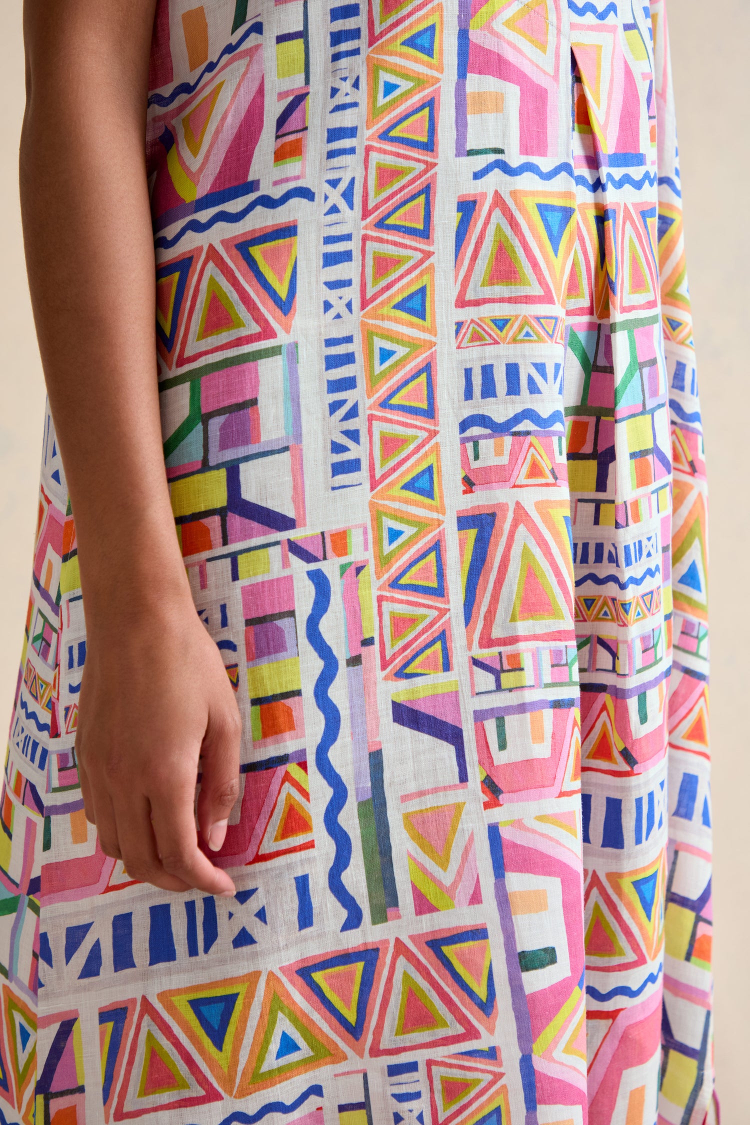 A person wears the Painted Geo Linen Dress, showcasing a vibrant geometric print with triangles, rectangles, and wavy lines in red, pink, green, blue, and yellow. Only the torso and arm are visible.