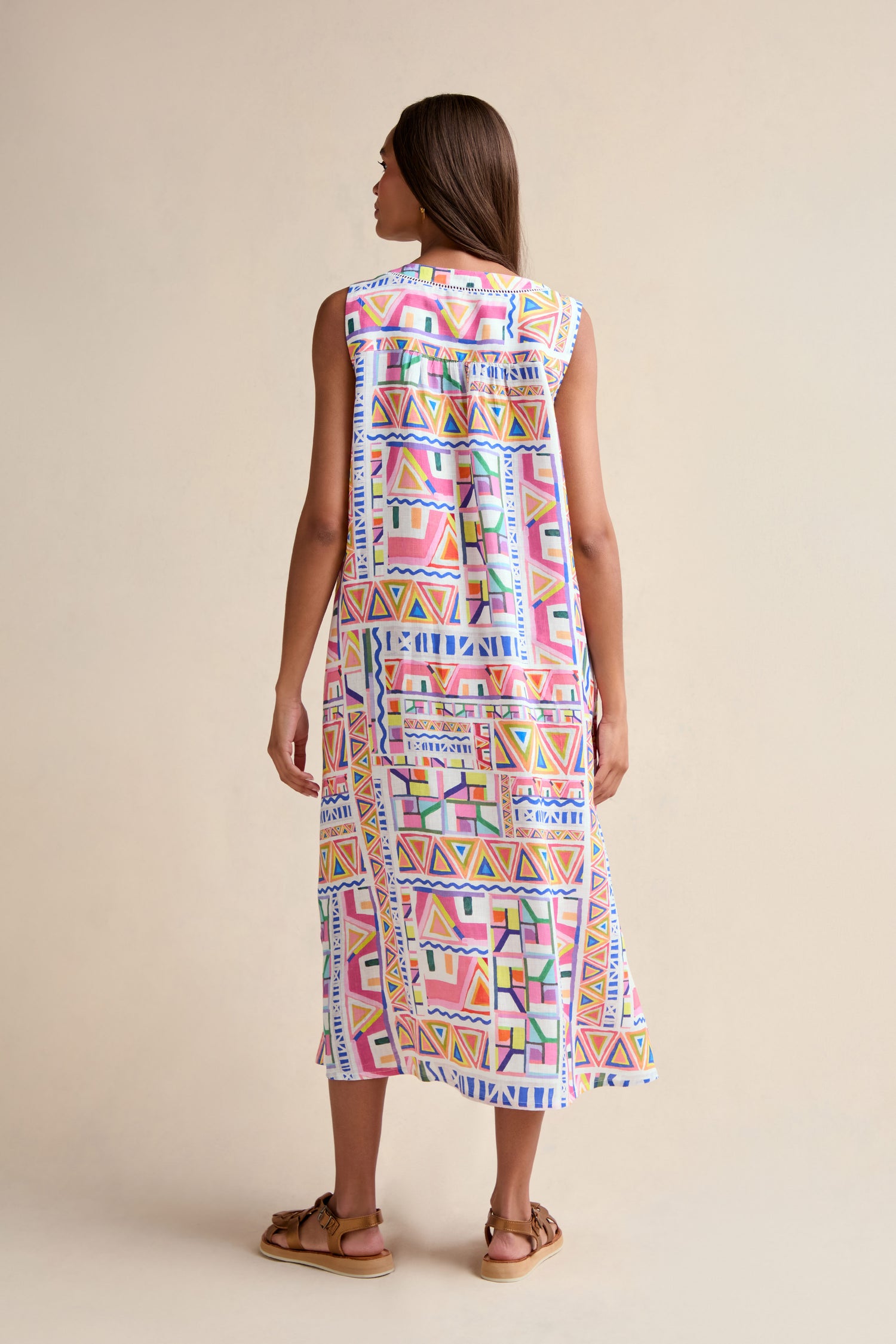 A woman faces away, wearing the Painted Geo Linen Dress—a sleeveless, midi-length linen dress with a colorful geometric print—paired with brown sandals against a plain light background.