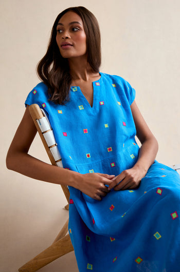 A woman sits in a wooden chair wearing the Embroidered Squares Linen Bubble Dress—a bright blue, short-sleeved, midi-length linen dress adorned with small, colorful square patterns.