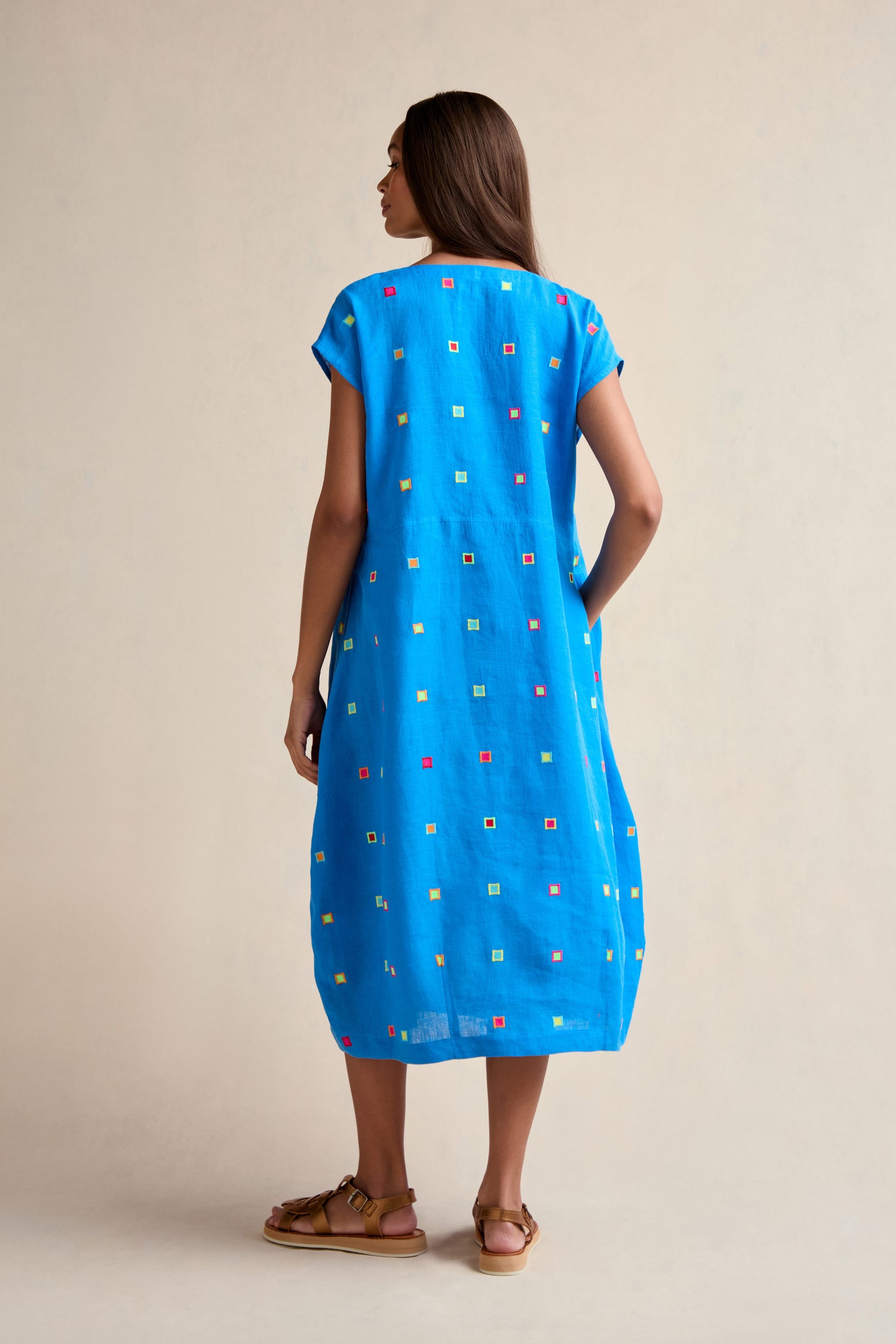 A person stands facing away, wearing the Embroidered Squares Linen Bubble Dress and brown sandals against a plain light background.