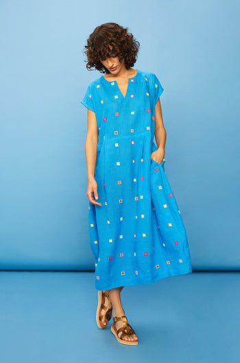 A woman in the Embroidered Squares Linen Bubble Dress—blue, midi-length, loose-fitting linen with colorful square embroidery and short sleeves—pairs it with brown sandals and stands against a blue background.