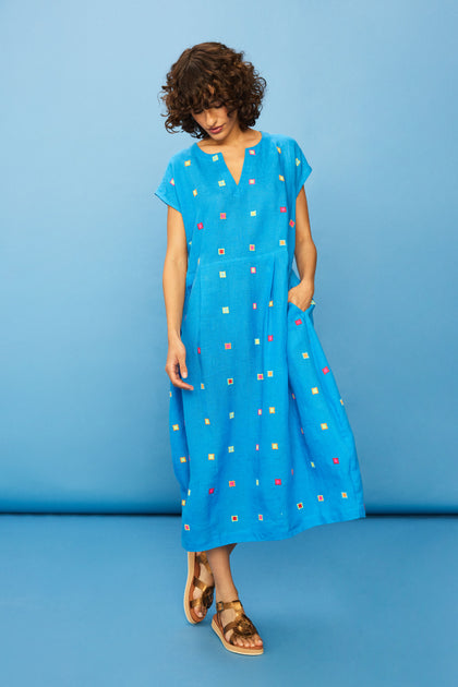 A woman in the Embroidered Squares Linen Bubble Dress—blue, midi-length, loose-fitting linen with colorful square embroidery and short sleeves—pairs it with brown sandals and stands against a blue background.