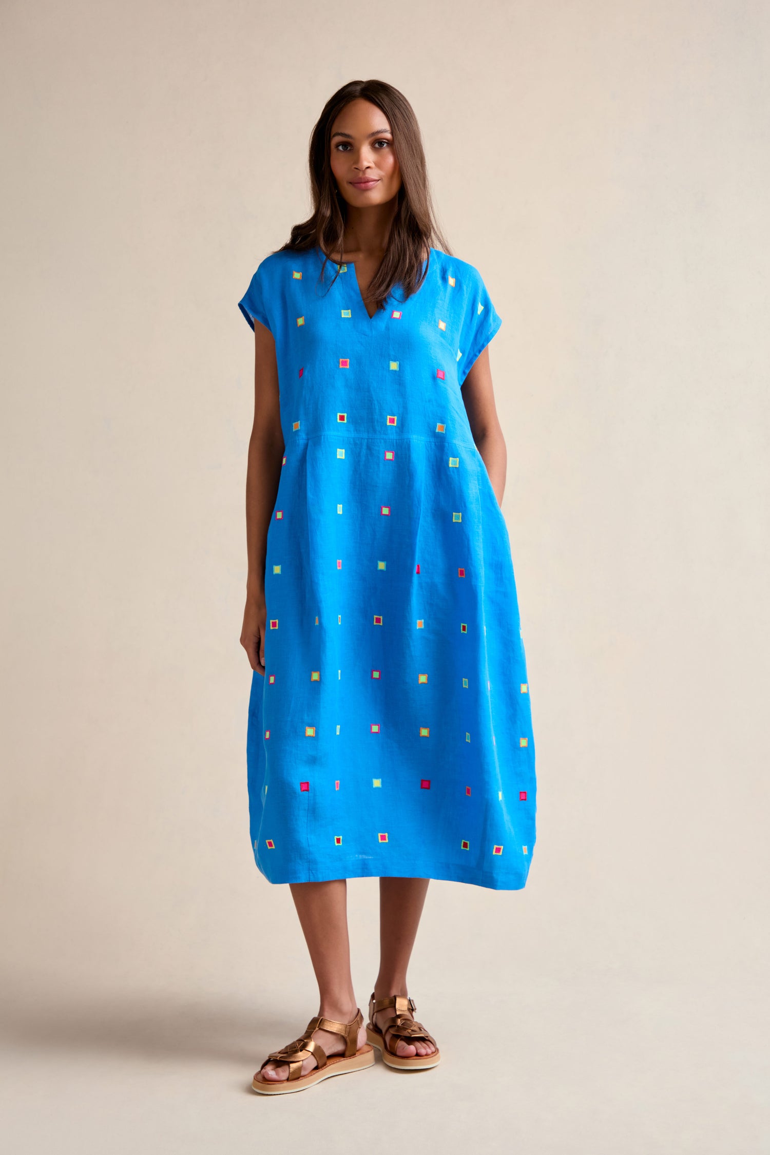 A woman with straight dark hair wears the Embroidered Squares Linen Bubble Dress—loose-fitting and midi-length with multicolored embroidery—paired with brown sandals, standing against a plain beige background.