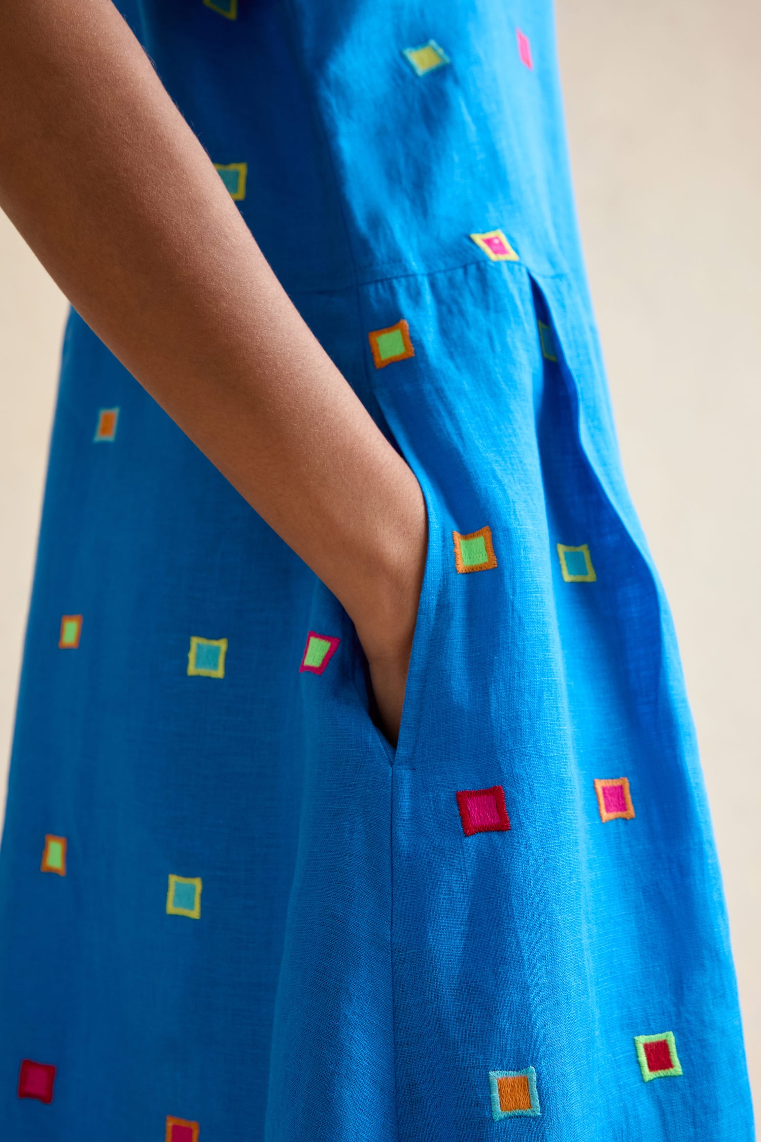 Close-up of a hand in the pocket of the Embroidered Squares Linen Bubble Dress, showcasing blue fabric with multicolored embroidery and a stylish midi-length design.