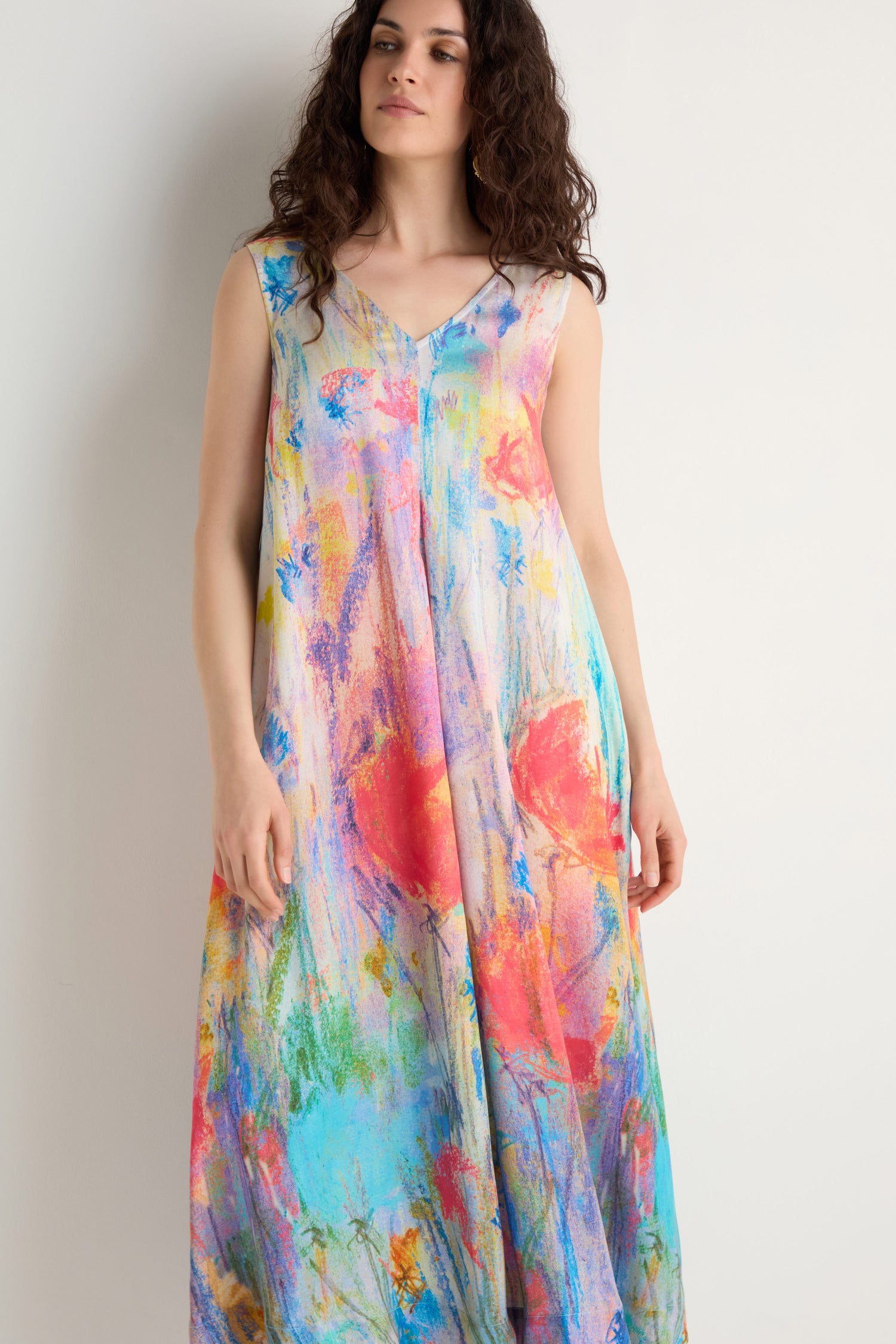 A woman stands against a white wall wearing the Meadow Dreams Sleeveless Dress, featuring a loose fit and a colorful, abstract wildflower print.