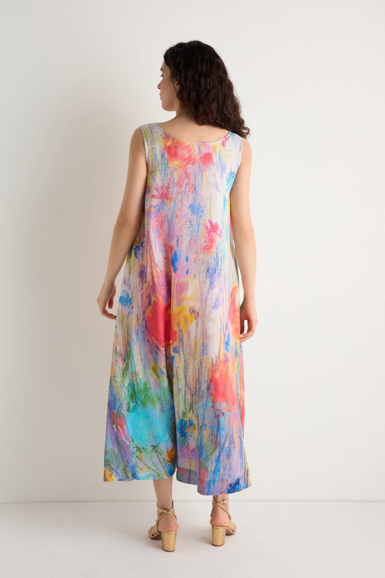 A woman, facing away, wears the Meadow Dreams Sleeveless Dress—an ankle-length, wildflower print A-line—with beige sandals against a white background.