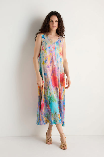 A woman stands against a white wall wearing the Meadow Dreams Sleeveless Dress, featuring a wildflower print, A-line silhouette, and paired with strappy sandals.
