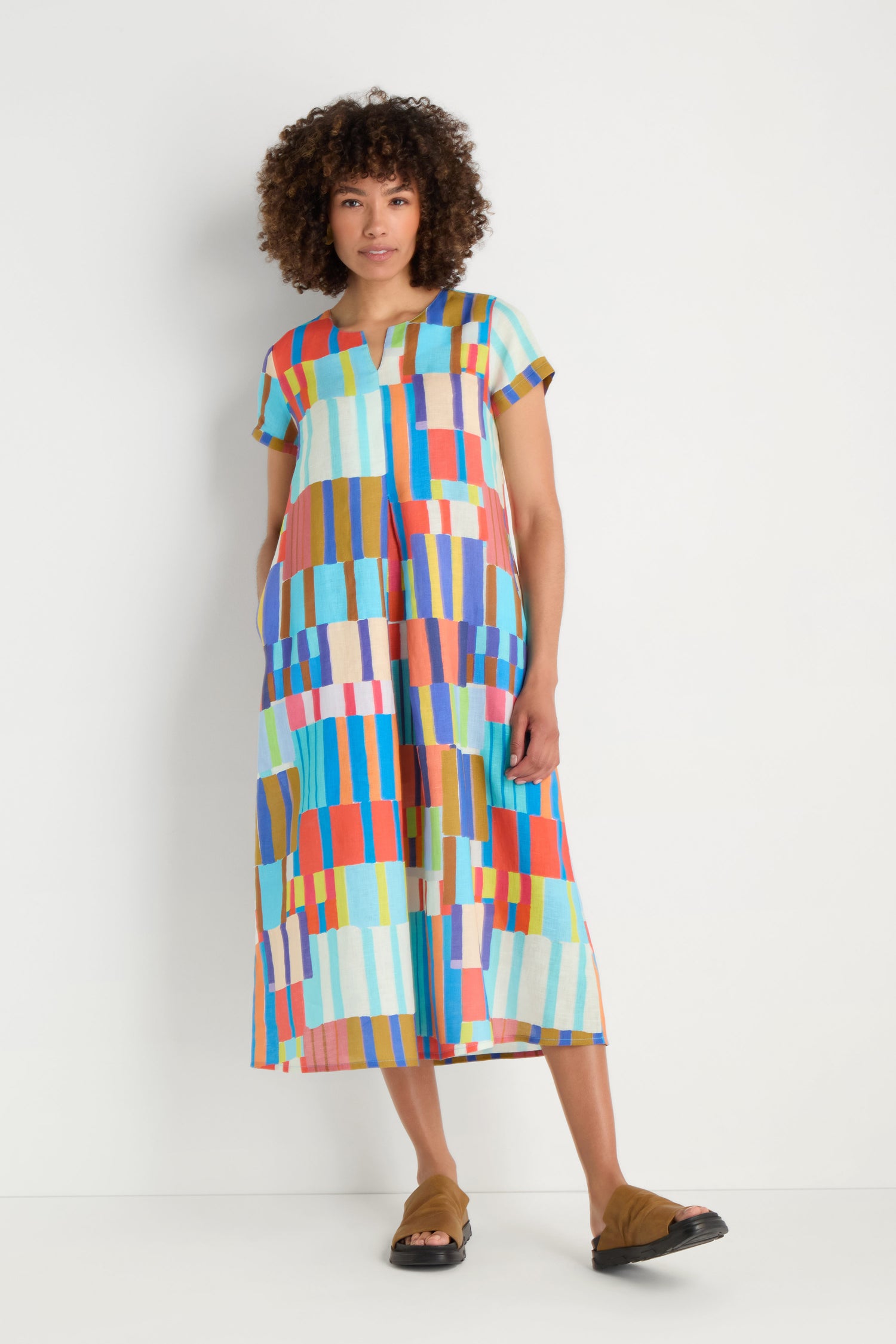 A person stands against a white background wearing the Painted Stripes A-Line Dress, an ankle-length linen dress with short sleeves and a vibrant geometric pattern, paired with brown sandals.