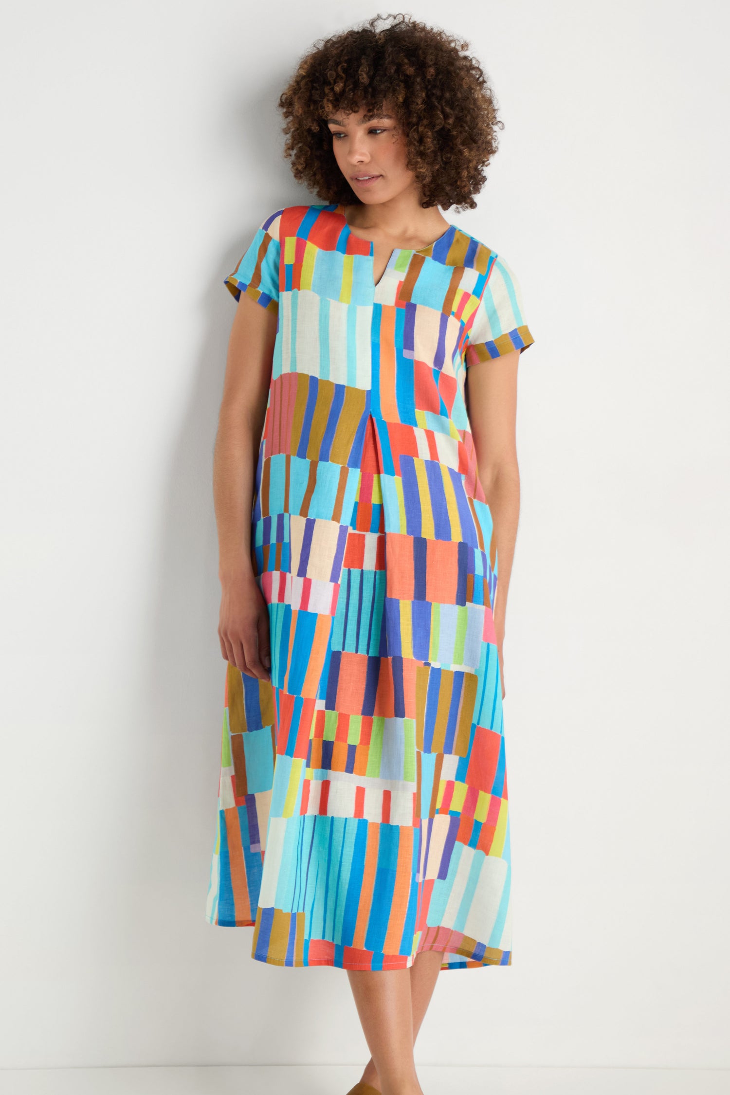 A woman stands against a white wall wearing the Painted Stripes A-Line Dress, featuring short sleeves and a bold multicolored geometric pattern.